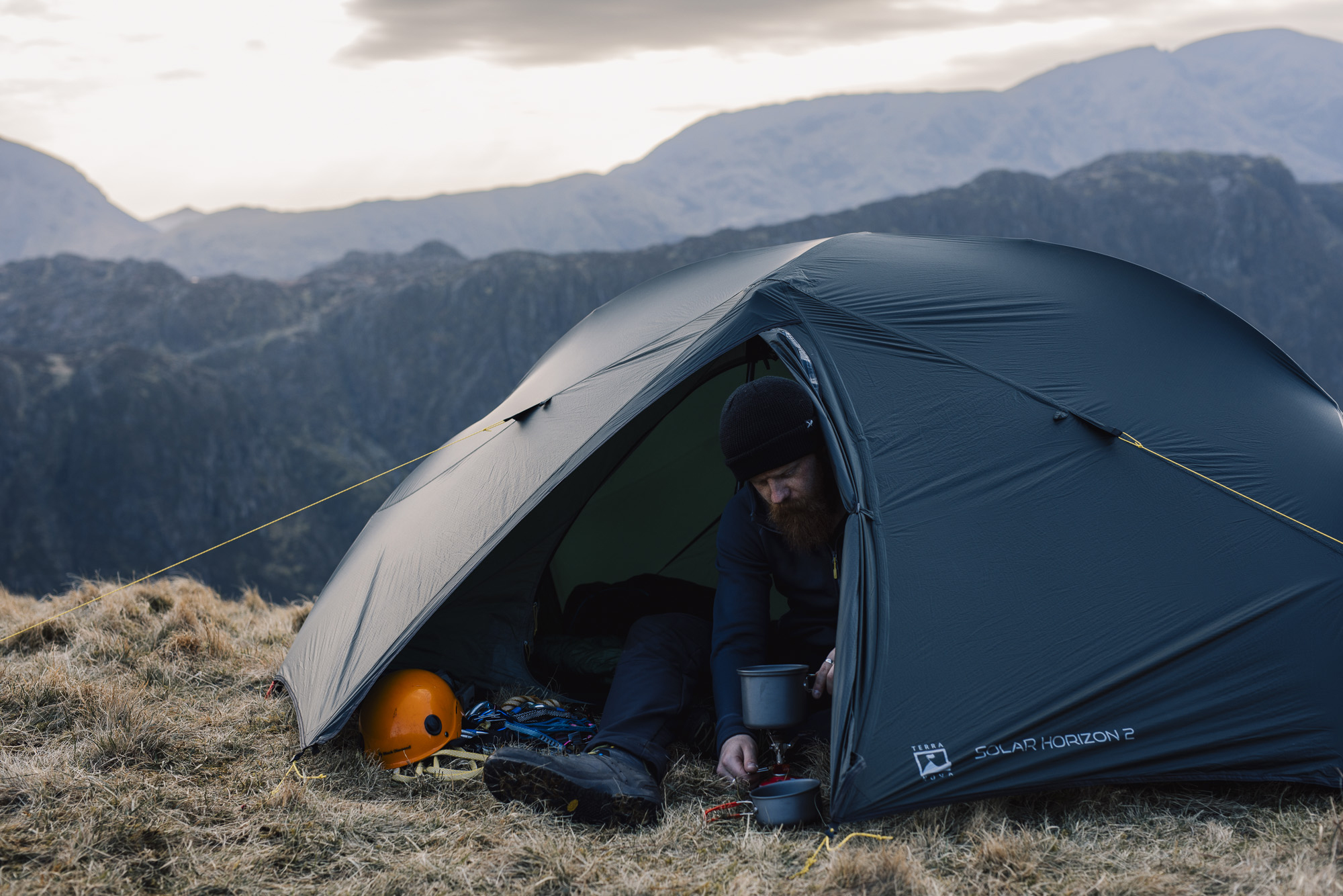 Bearded man in a black beanie crouches at the open door of a dark two-person tent on a grassy mountain ridge, preparing a hot drink on a small camping stove beside an orange climbing helmet and ropes, with layered rocky peaks in the misty background.