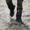 Tay-Ankle-GORE-TEX-Gaiter-17-LR-3.jpg Lower legs wearing brown trousers and black waterproof gaiters with an orange tab and hiking boots splashing through a shallow rocky stream.