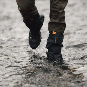 Lower legs wearing brown trousers and black waterproof gaiters with an orange tab and hiking boots splashing through a shallow rocky stream.