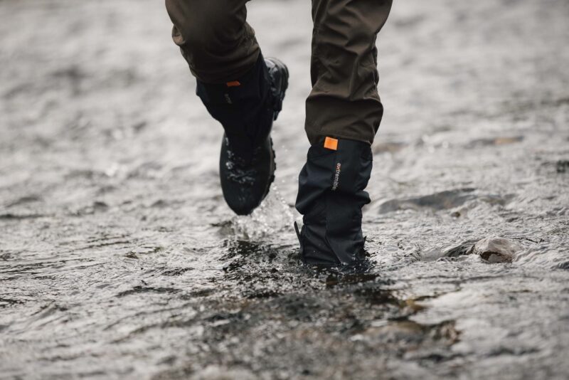 Tay-Ankle-GORE-TEX-Gaiter-17-LR-3.jpg Lower legs wearing brown trousers and black waterproof gaiters with an orange tab and hiking boots splashing through a shallow rocky stream.
