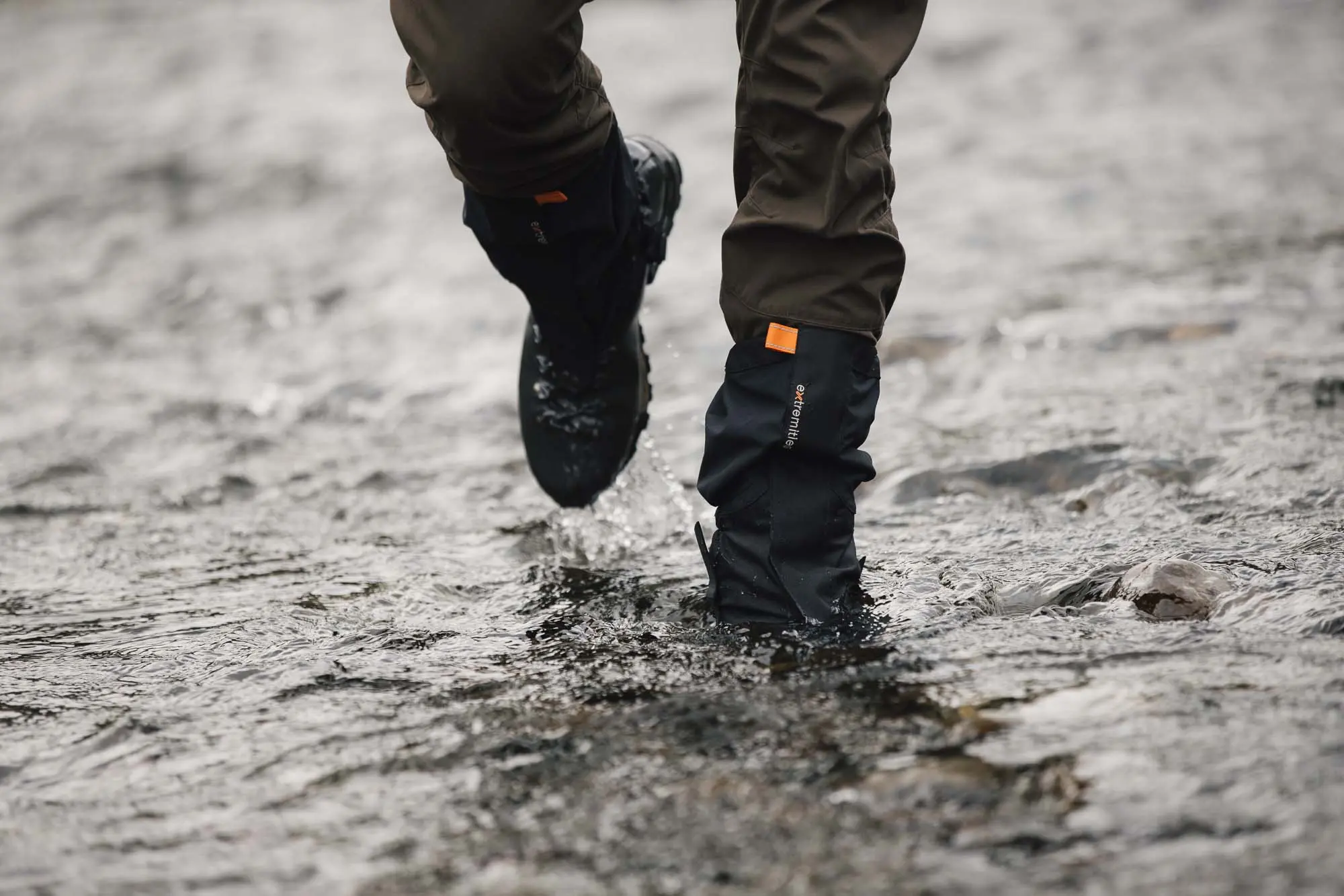 Lower legs wearing brown trousers and black waterproof gaiters with an orange tab and hiking boots splashing through a shallow rocky stream.