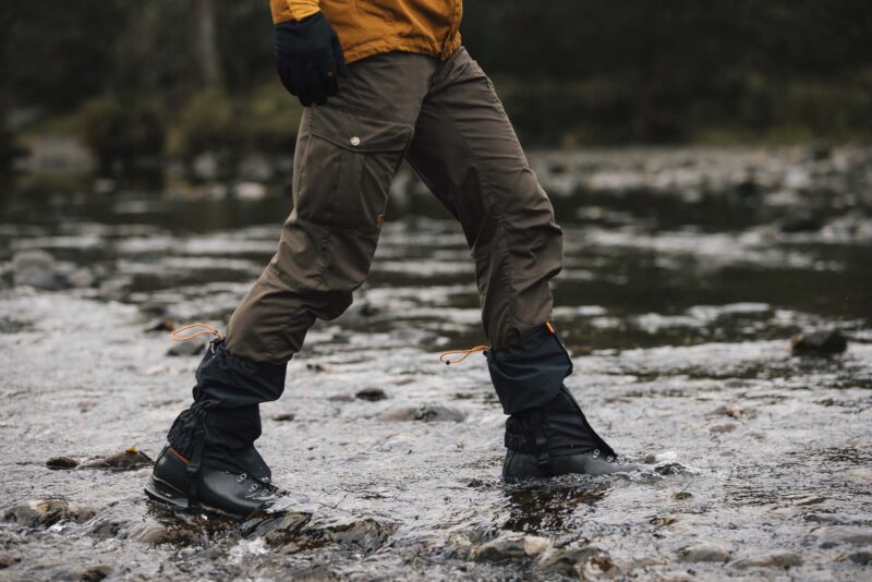 Tay-Ankle-GORE-TEX-Gaiter-24-LR-3.jpg Person's lower body in brown cargo trousers, black gaiters with orange drawcords and hiking boots stepping across a shallow rocky stream with water splashing around the boots