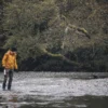 Tay-Ankle-GORE-TEX-Gaiter-8-LR-3.jpg Person in a yellow jacket, black beanie and waders walking through a shallow, pebble-strewn river holding a metal water bottle, with low, mossy trees overhanging the bank in the background.