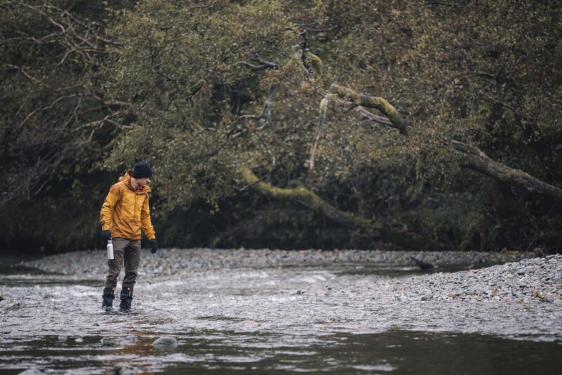 Tay-Ankle-GORE-TEX-Gaiter-8-LR-3.jpg Person in a yellow jacket, black beanie and waders walking through a shallow, pebble-strewn river holding a metal water bottle, with low, mossy trees overhanging the bank in the background.