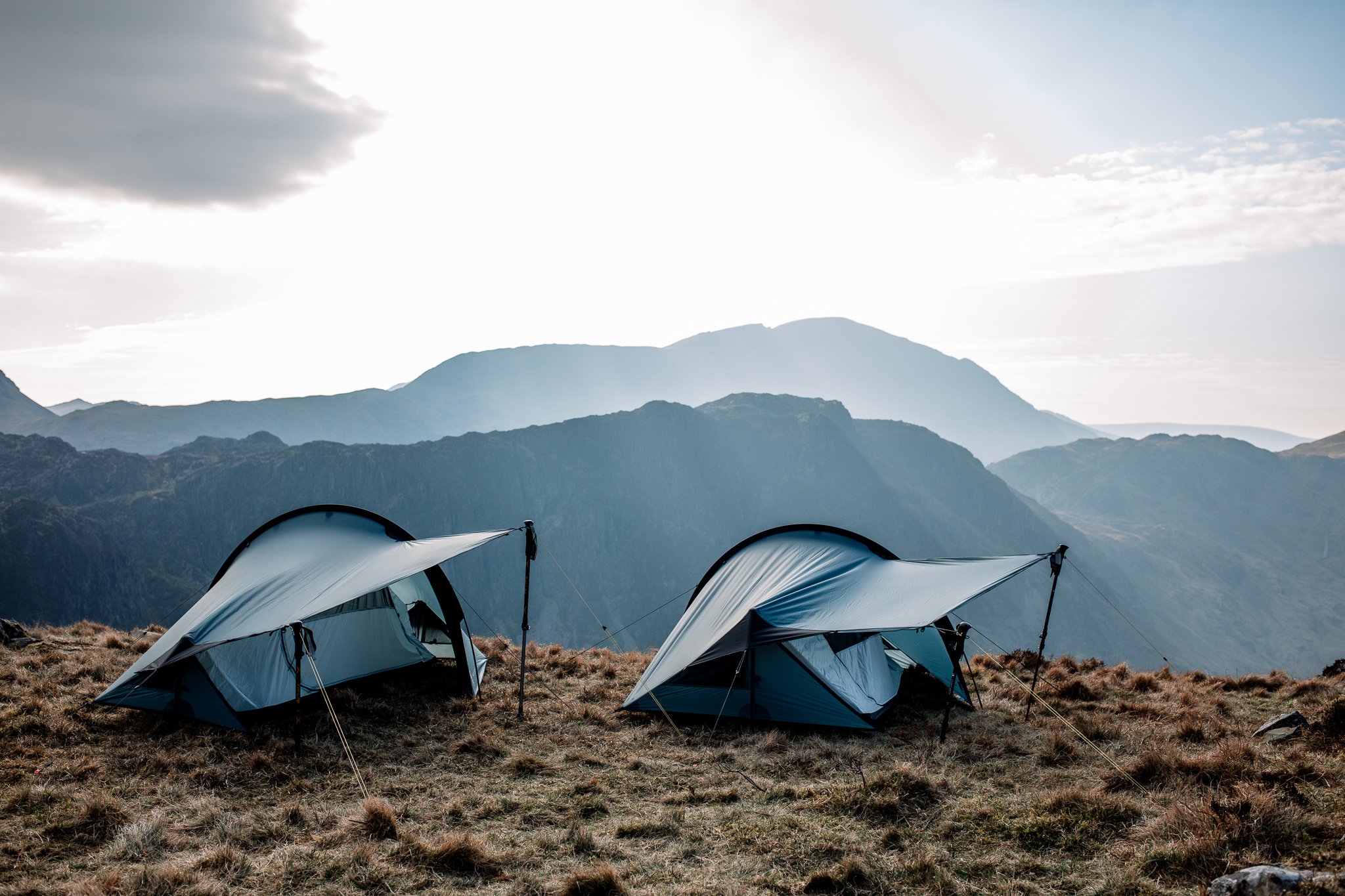 Terra-Nova-Halny-1-2-LS2-3.webp Two blue backpacking tents with extended vestibules held by trekking poles pitched on a grassy mountain plateau, backlit by a bright sky and layered hazy mountain ridges in the distance.