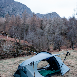 A small light-grey backpacking tent propped open by trekking poles on a grassy hillside, a person in dark clothing arranging a green sleeping bag inside with bare trees and rocky mountains under a pale sky in the background.