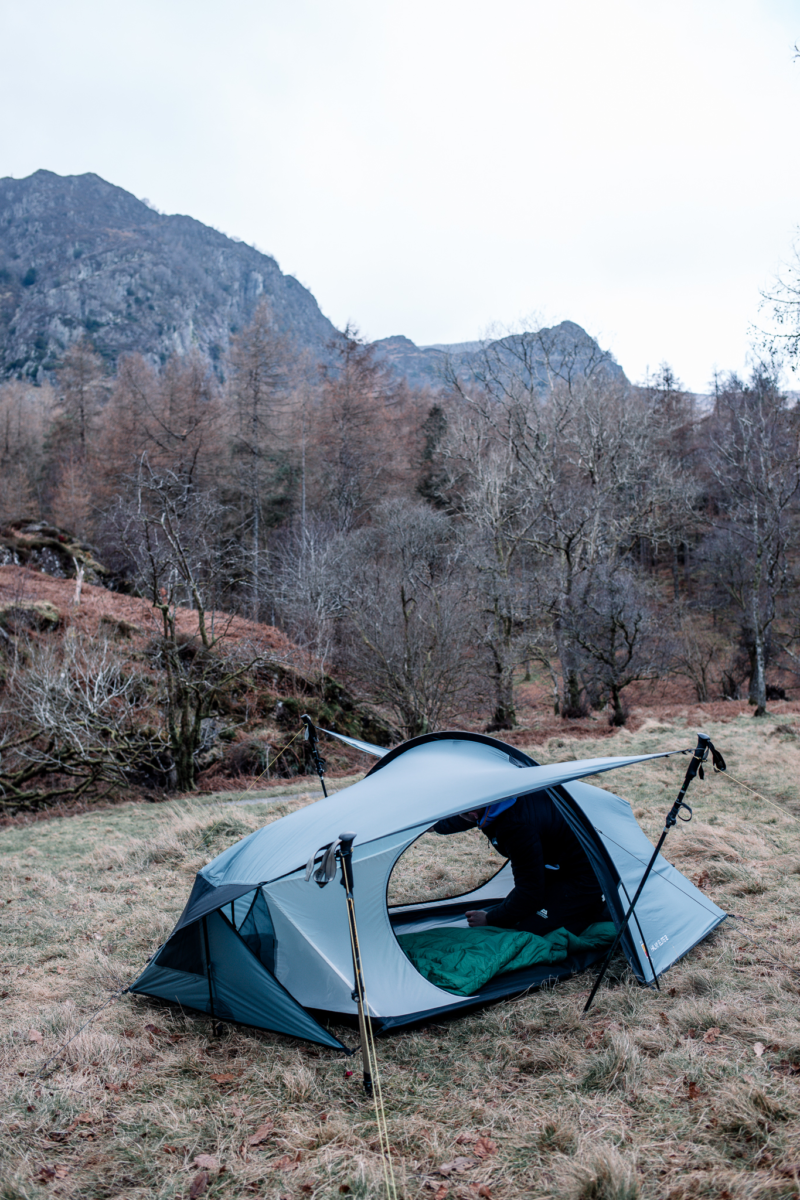 A small light-grey backpacking tent propped open by trekking poles on a grassy hillside, a person in dark clothing arranging a green sleeping bag inside with bare trees and rocky mountains under a pale sky in the background.