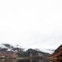 Green camping tent pitched on a grassy lakeshore with misty, rugged hills reflected in the calm water under an overcast sky