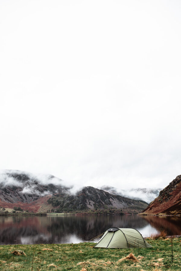 Green camping tent pitched on a grassy lakeshore with misty, rugged hills reflected in the calm water under an overcast sky