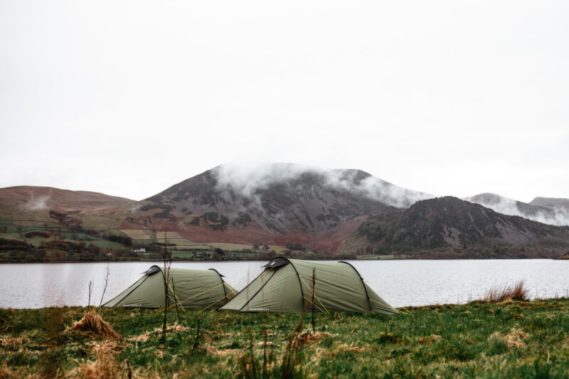 Two green tents pitched on a grassy lakeshore with a calm lake and mist-shrouded hills under an overcast sky.