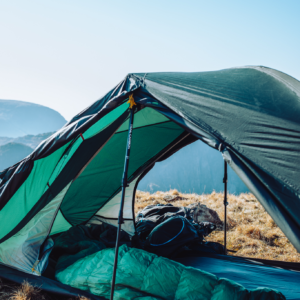 Green two-person tent propped open with trekking poles on a dry grassy mountain ridge, showing a green sleeping bag and backpack inside and distant blue mountains under a clear sky.