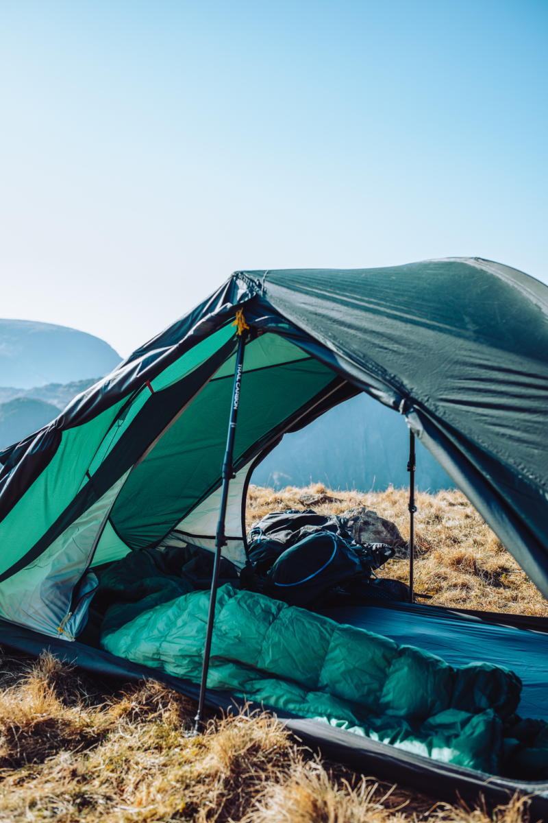 Green two-person tent propped open with trekking poles on a dry grassy mountain ridge, showing a green sleeping bag and backpack inside and distant blue mountains under a clear sky.