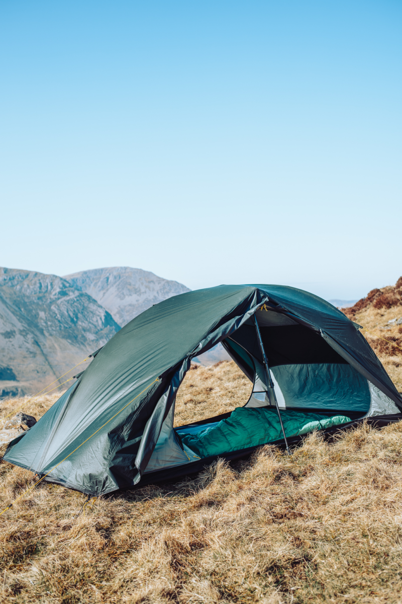 Green two-person dome tent pitched on a dry grassy mountain slope with its front flap open revealing a green sleeping bag inside and distant rocky peaks under a clear blue sky.