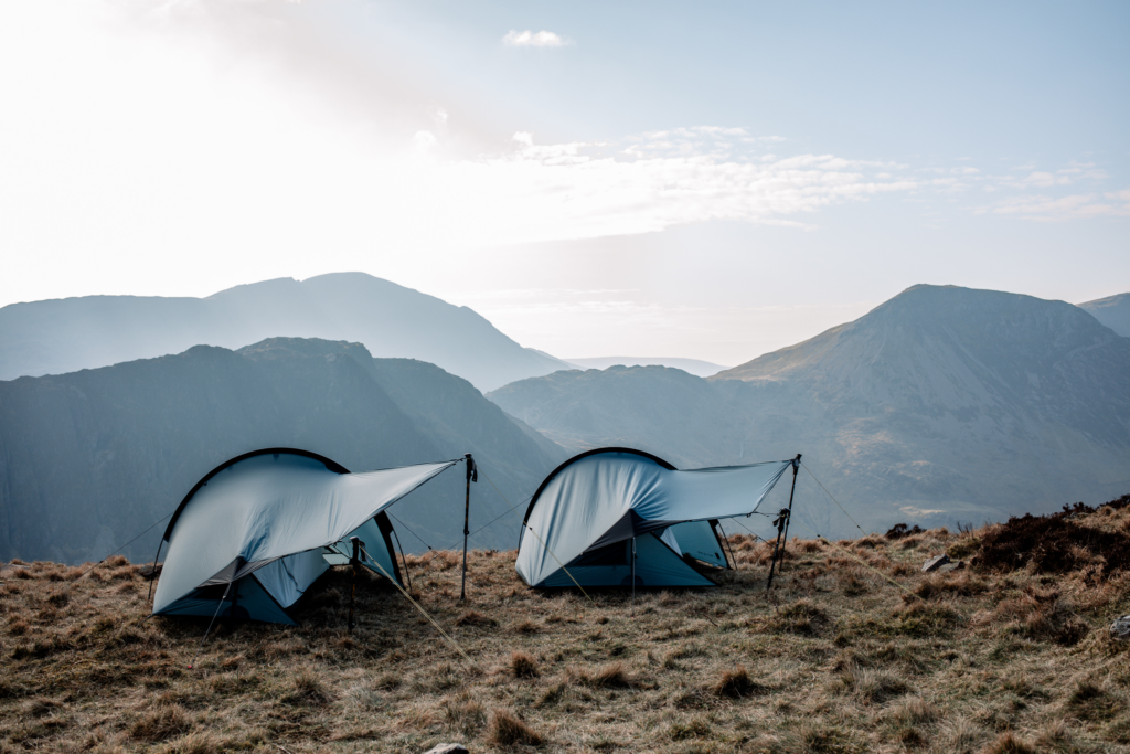 Terra-Nova-halny-1-2-LS1-3.webp Two low-profile blue-grey tents staked on a windswept grassy ridge, their curved rainflies propped with trekking poles and guylines, with layered misty mountains and a pale morning sky in the background.