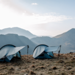 Two low-profile blue-grey tents staked on a windswept grassy ridge, their curved rainflies propped with trekking poles and guylines, with layered misty mountains and a pale morning sky in the background.