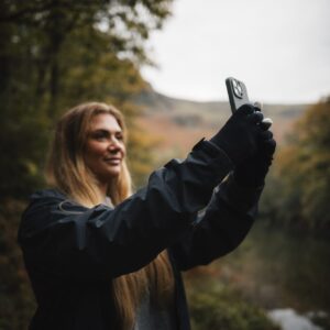 A woman in a dark rain jacket and gloves holds up a smartphone to take a photo, smiling against an overcast autumn woodland backdrop with a river and distant hills.