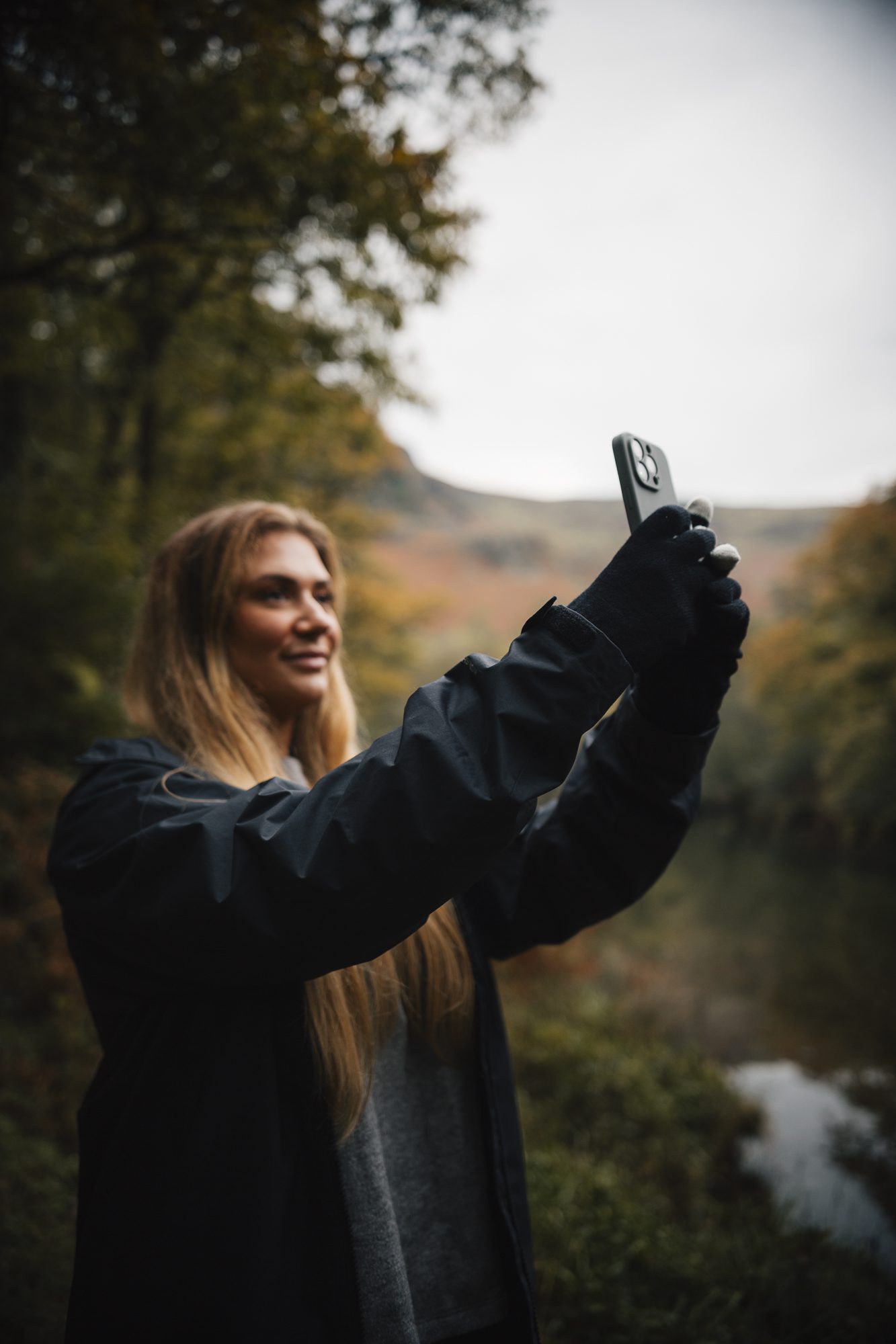 A woman in a dark rain jacket and gloves holds up a smartphone to take a photo, smiling against an overcast autumn woodland backdrop with a river and distant hills.