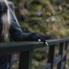 Thinny-Touch-LRR-3.jpg Partial view of a person with long blonde hair in a black coat, resting a black-gloved hand with white fingertips on a dark wooden bridge railing against an out-of-focus autumnal woodland and a nearby branch with yellowing leaves.