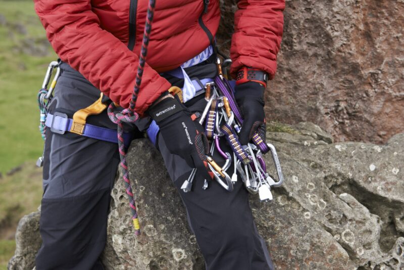 Tor-Glove-lifestyle-photo-scaled-2.jpg Climber's torso and gloved hands in a red jacket seated on a rock, holding a cluster of silver carabiners, colourful slings and quickdraws clipped to a blue-and-yellow harness with a braided rope running through.