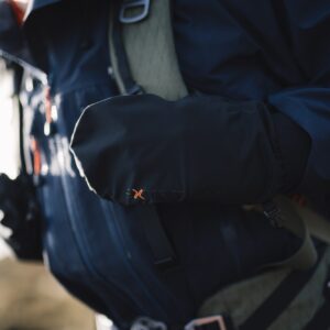 Close-up of a hiker wearing a dark Gore‑Tex jacket with a black insulated mitten bearing an orange 'x' logo resting over a backpack strap