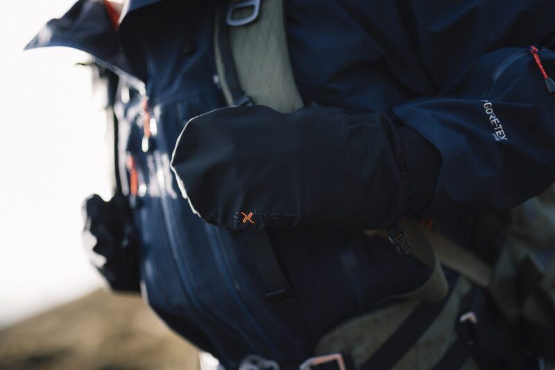Torrent-Mitt-10-3.jpg Close-up of a hiker wearing a dark Gore‑Tex jacket with a black insulated mitten bearing an orange 'x' logo resting over a backpack strap