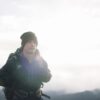 Torrent-Mitt-16-3.jpg Hiker in a dark beanie and insulated jacket carrying a backpack, standing on a ridge with a bright overcast sky, distant mountains and a faint lens flare across his chest.