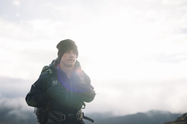 Torrent-Mitt-16-3.jpg Hiker in a dark beanie and insulated jacket carrying a backpack, standing on a ridge with a bright overcast sky, distant mountains and a faint lens flare across his chest.