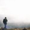 Torrent-Mitt-3-3.jpg A lone hiker in a black beanie and dark jacket carrying a large backpack walks across grassy, fog‑shrouded mountain terrain under a bright, overcast sky.
