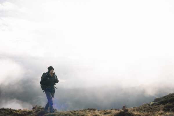 Torrent-Mitt-3-3.jpg A lone hiker in a black beanie and dark jacket carrying a large backpack walks across grassy, fog‑shrouded mountain terrain under a bright, overcast sky.