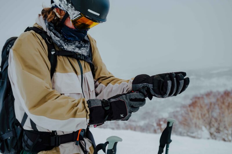 Torres-Peak-3-scaled-4.jpg Skier in a beige-and-white jacket wearing a black helmet, yellow goggles and a snow-speckled neck gaiter adjusts grey-and-black gloves next to ski poles on a snowy slope.