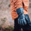 Torres_Peak_LS-2-scaled-4.jpg Close-up of a person in an orange jacket showing a grey insulated glove with an adjustable black strap, set against a blurred brown-green outdoor background.