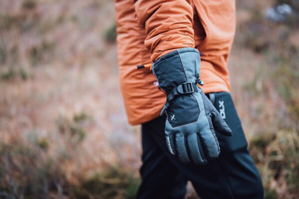 Torres_Peak_LS-2-scaled-4.jpg Close-up of a person in an orange jacket showing a grey insulated glove with an adjustable black strap, set against a blurred brown-green outdoor background.