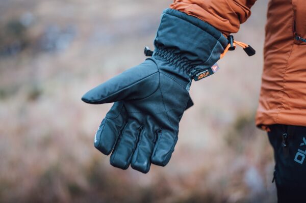 Torres_Peak_LS-5-scaled-4.jpg Close-up of an outstretched black insulated winter glove with an orange drawcord and small logo, worn by a person in an orange jacket against a soft-focus outdoor background.