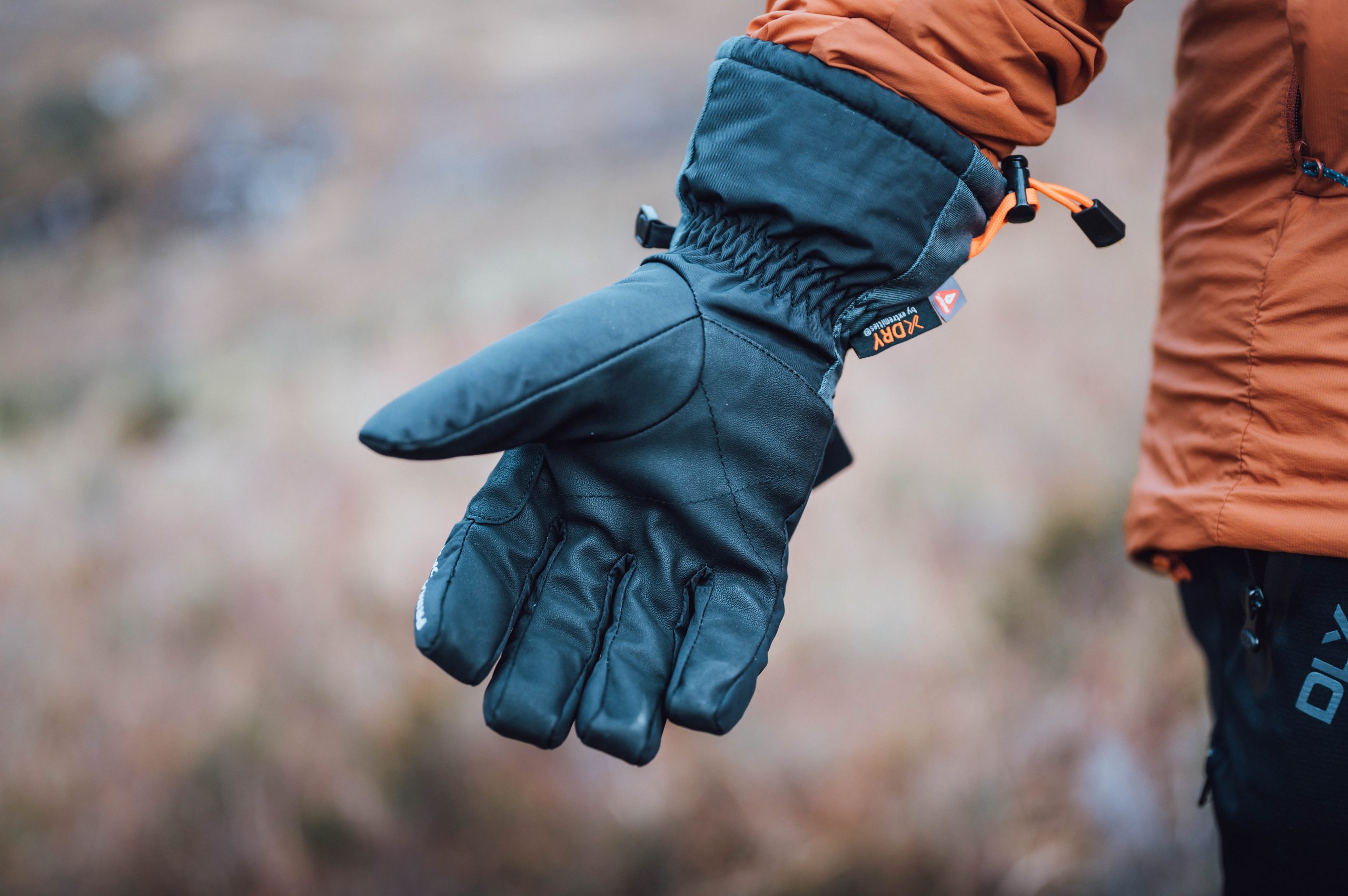 Close-up of an outstretched black insulated winter glove with an orange drawcord and small logo, worn by a person in an orange jacket against a soft-focus outdoor background.