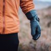 Torres_Peak_LS-7-scaled-4.jpg Close-up of a person wearing an orange jacket with their arm at their side in a dark insulated glove with a buckle strap, set against a blurred grassy outdoor background.
