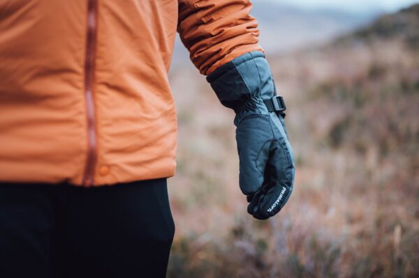 Torres_Peak_LS-7-scaled-4.jpg Close-up of a person wearing an orange jacket with their arm at their side in a dark insulated glove with a buckle strap, set against a blurred grassy outdoor background.