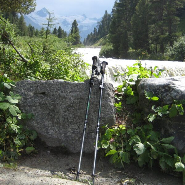 Trail-Elite-Trekking-Pole-Lifestyle-Square-1500x1500-1-3.jpg Two trekking poles leaning against a large granite boulder beside a fast-flowing mountain stream, surrounded by green foliage and pine trees with snow-capped peaks visible in the distance.