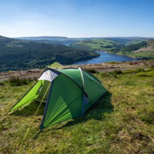 Green backpacking tent pitched on a grassy hilltop overlooking a blue reservoir with a dam and forested, rolling hills under a clear blue sky.