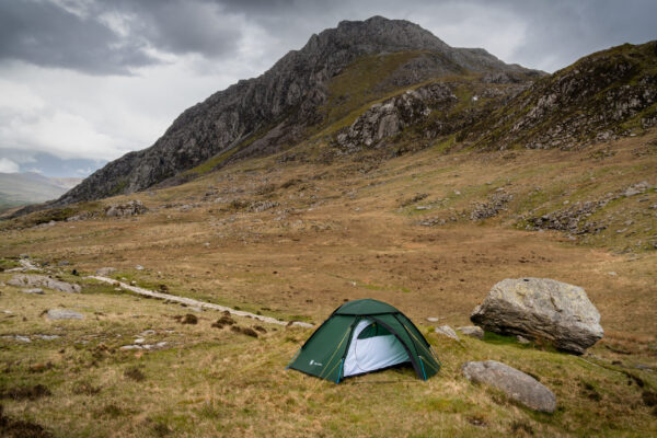 Terrs Nova Snowdonia Photography Shoot May 2023 Green dome tent pitched on grassy moor beside a large boulder in a broad, rocky mountain valley with steep crags rising in the background under an overcast sky.