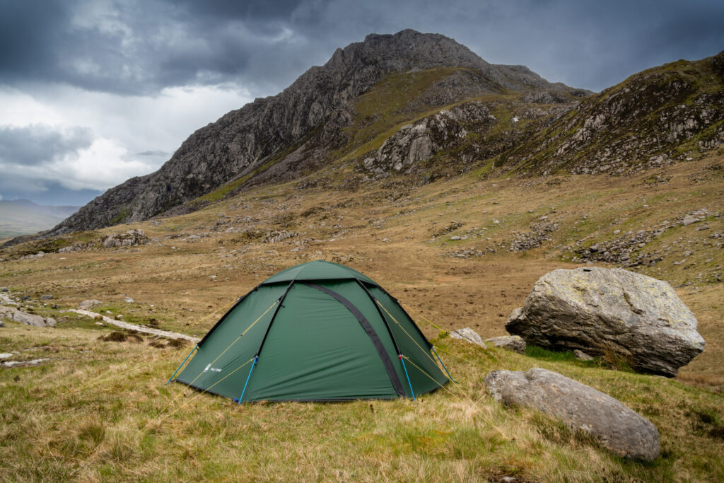 Green dome tent anchored with yellow guylines and blue poles pitched on grassy moorland beside large boulders, with steep rocky mountains rising in the background under a dark, stormy sky.