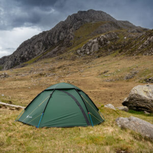 Green dome tent anchored with yellow guylines and blue poles pitched on grassy moorland beside large boulders, with steep rocky mountains rising in the background under a dark, stormy sky.