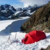 Ven.SG10.Camp7_-scaled-4.jpg A bright red mountaineering tent pitched in deep snow beside a rocky ridge with skis and poles propped nearby, set against a sweeping glacier and jagged snow-covered mountains under a blue sky.