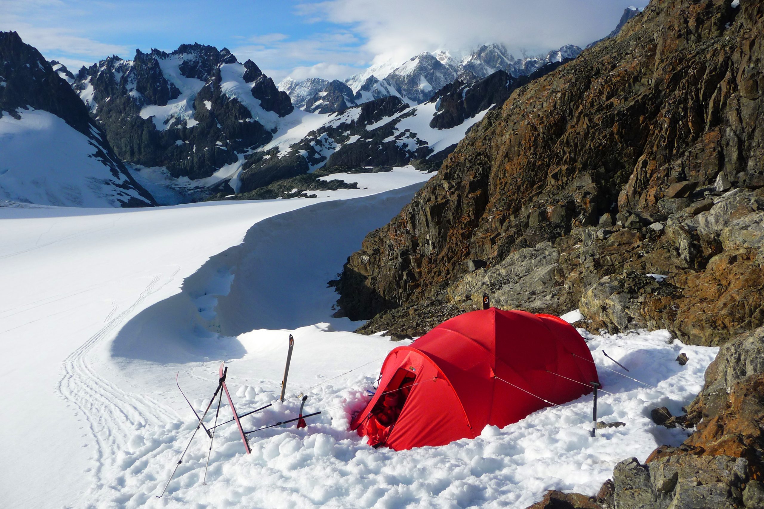 A bright red mountaineering tent pitched in deep snow beside a rocky ridge with skis and poles propped nearby, set against a sweeping glacier and jagged snow-covered mountains under a blue sky.