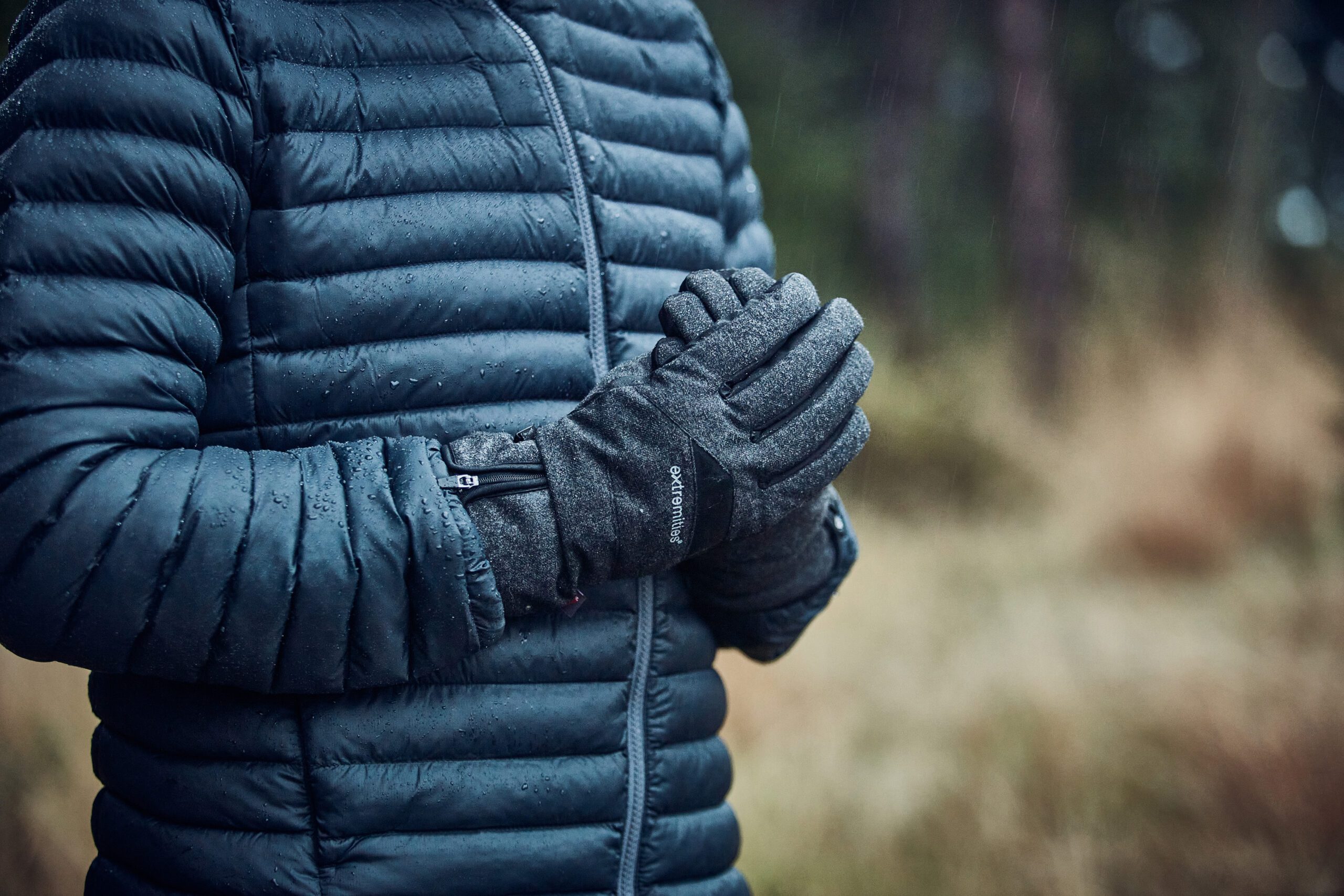 Vermont-Gloves-scaled-2.jpg Torso of a person in a dark blue quilted puffer jacket with raindrops, hands clasped in grey insulated gloves against a blurred rainy woodland background.