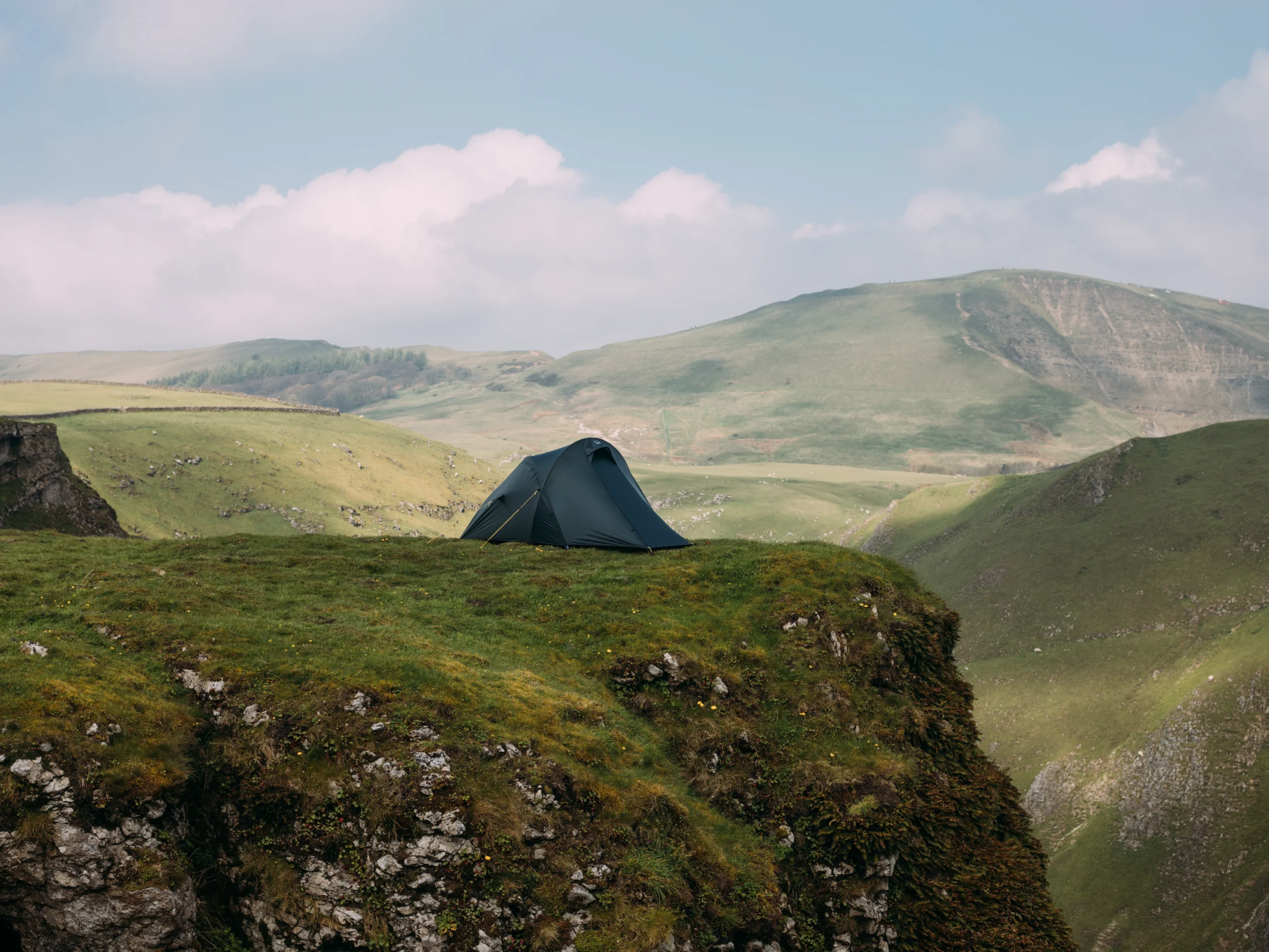 Camper in a blue HELM ELITE 1 tent sits at the open entrance wearing a mustard jacket and handling a small tool, surrounded by grassy tussocks with hazy mountains in the background.