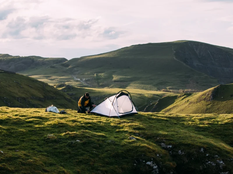 A camper kneeling to pitch a small white tent on a grassy ridge with rolling green hills and a cloudy sky in the background.