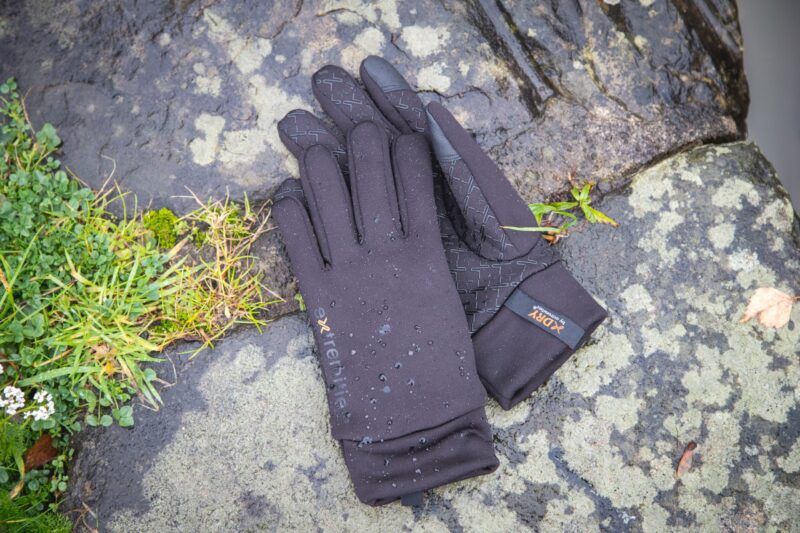 A pair of black waterproof gloves with textured grip patterns and water droplets lying on wet, lichen-covered rocks beside a small patch of grass.
