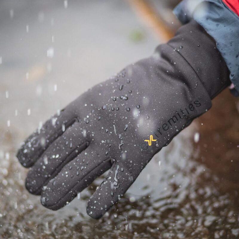 Close-up of a black waterproof glove covered in beaded rain droplets with a small yellow logo and text on the wrist, hand held over a rippling puddle as rain falls.