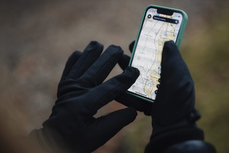 Waterproof-Power-Liner-Gloves-1-3.jpg Hands in black gloves holding a smartphone with a green case displaying a detailed topographic map, set against a blurred outdoor background.