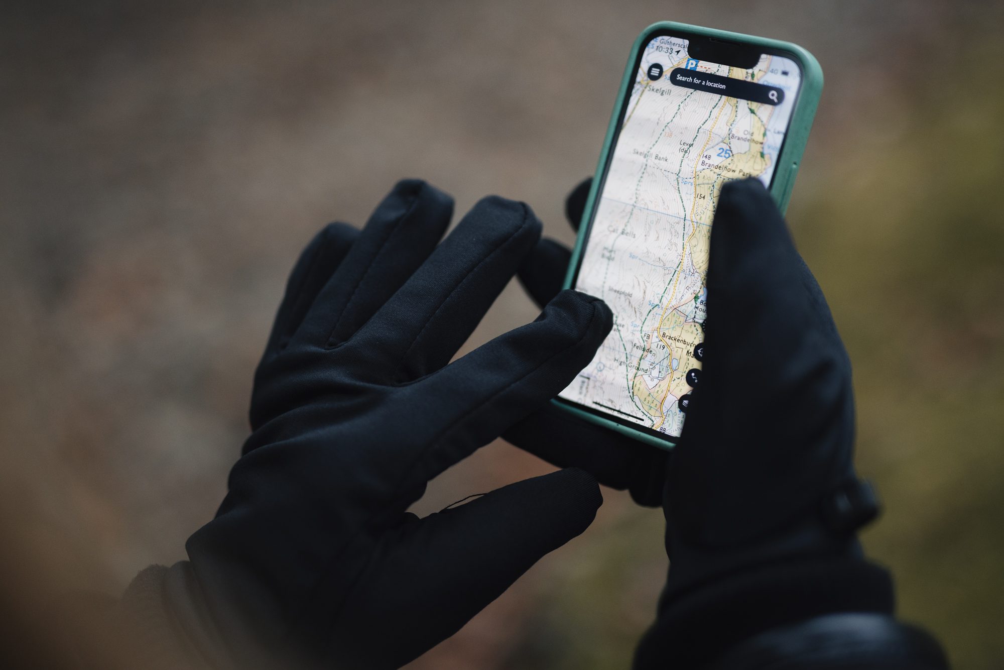 Hands in black gloves holding a smartphone with a green case displaying a detailed topographic map, set against a blurred outdoor background.